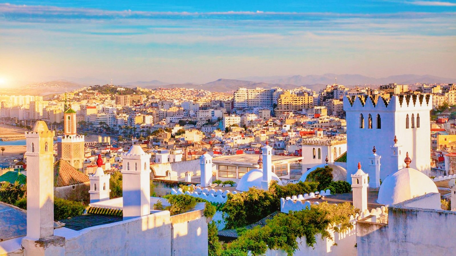 Sunlit cityscape with white buildings and blue-domed towers along a hillside at sunset.