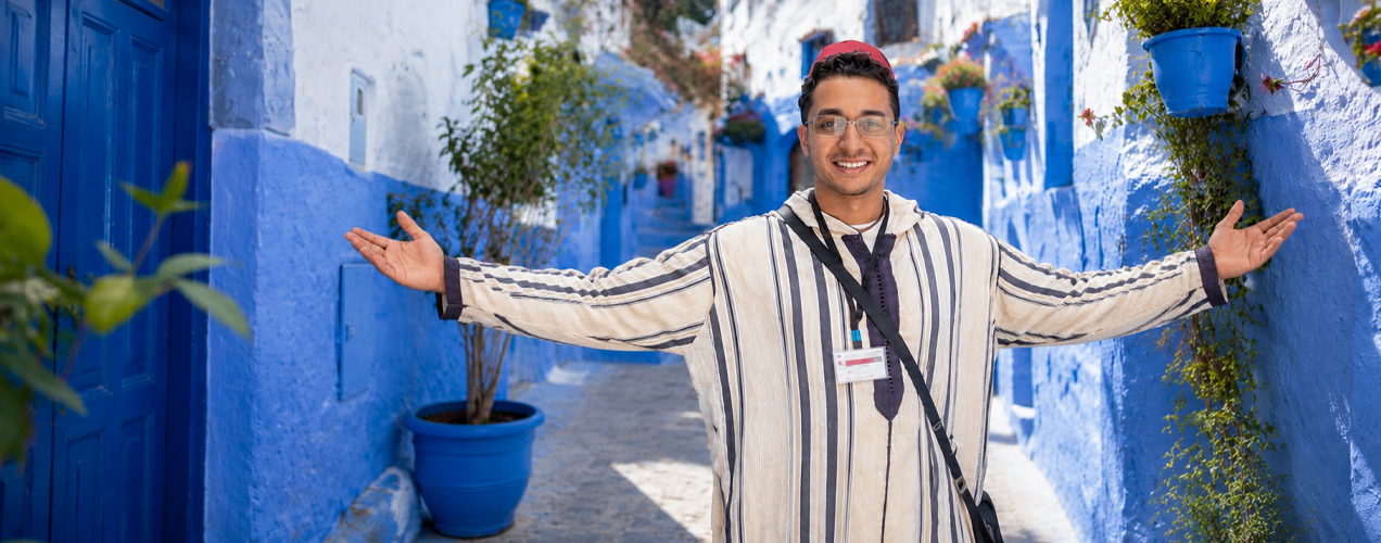 Smiling man in a striped traditional robe with arms outstretched in a sunlit blue-washed alley lined with blue walls and potted plants.
