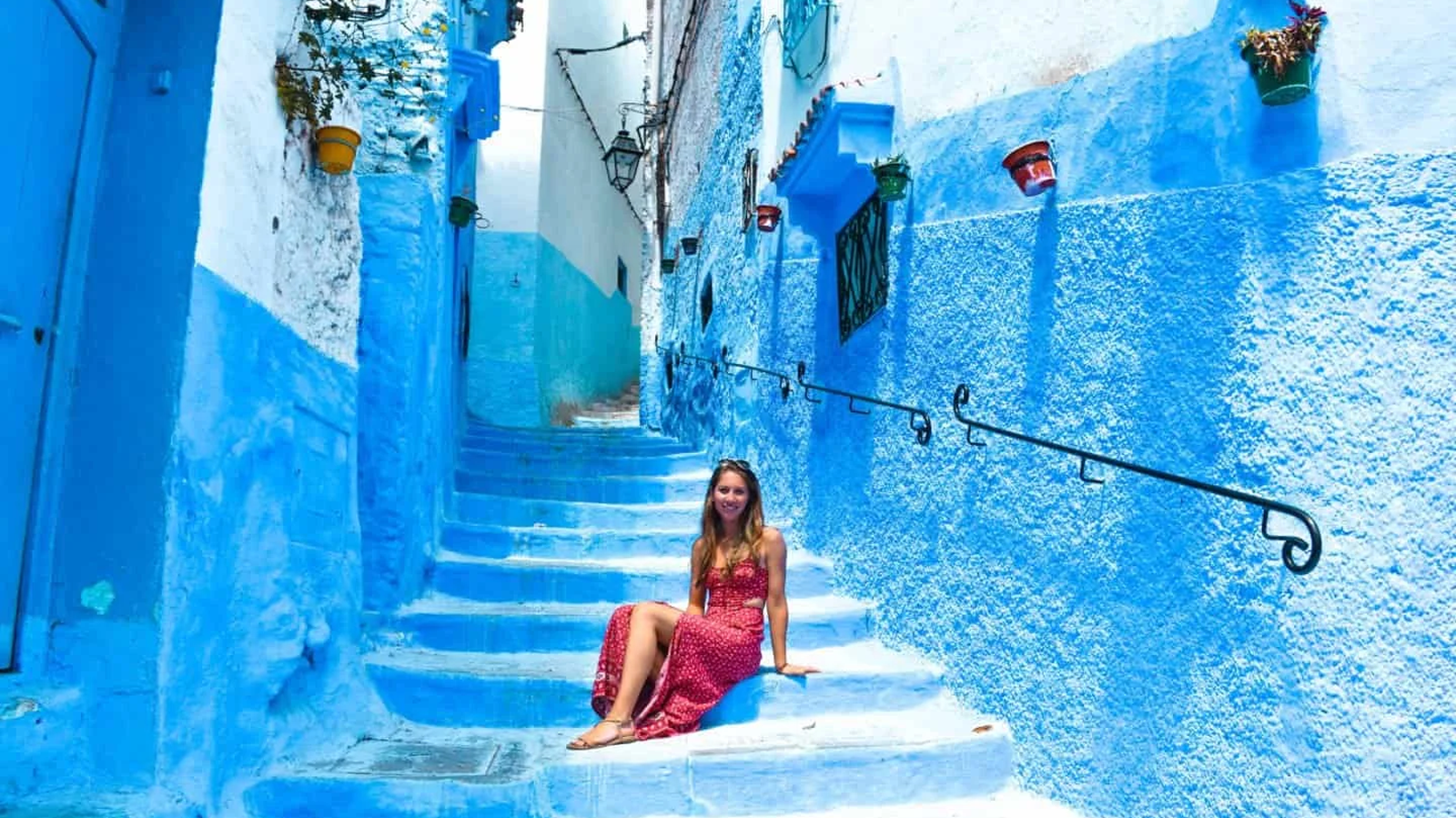 Woman in a red dress sits on blue-painted stairs in a narrow blue alley.