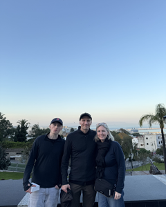 Three people posing on a terrace overlooking the coast, with palm trees and buildings in the background, under a clear blue sky there is the ocean.