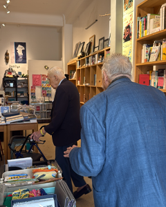 Two elderly men browse shelves in a cozy bookstore, surrounded by stacks of books, magazines, and posters on the walls.