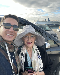 Two smiling people posing for a selfie beside a parked car, with a waterfront city skyline in the background; man wears sunglasses and a plaid scarf, woman in a wide-brim hat and scarf holding a pink phone.