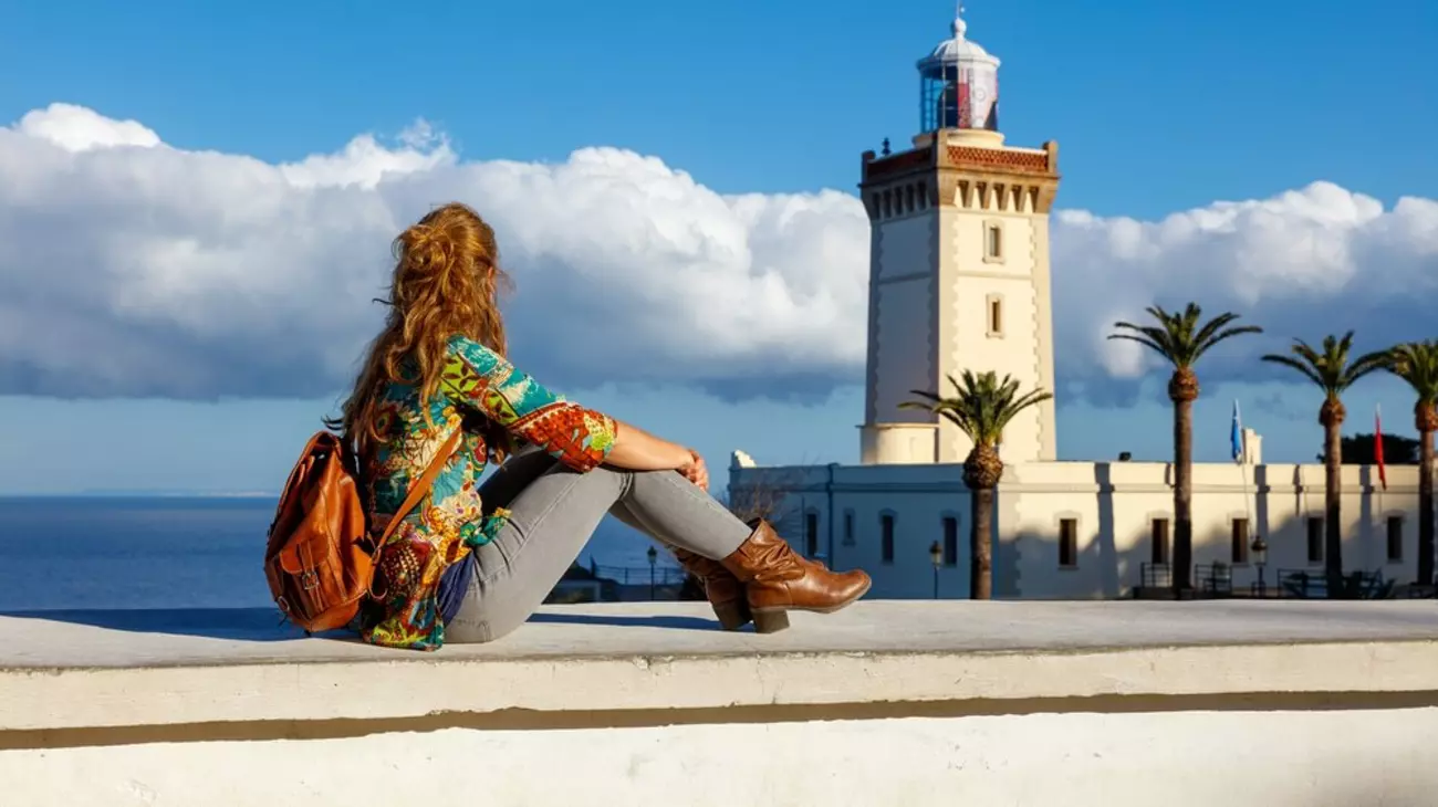 Person sitting on a sea wall with a brown backpack, overlooking a white lighthouse and palm trees by the sea.